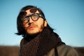 Outdoors, close-up portrait of young serious man with long hair wearing hipster round sunglasses and scarf. Blue sky background Royalty Free Stock Photo