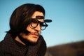 Outdoors, close-up portrait of young thoughtful man with long hair wearing hipster round sunglasses and scarf. Blue sky background Royalty Free Stock Photo