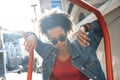 Outdoors Leisure. Young stylish african girl in denim jacket and sunglasses standing leaning on bar cool in sunlight Royalty Free Stock Photo