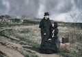 Outdoors portrait of a victorian lady in black sitting on the road with her luggage and gentleman standing nearby