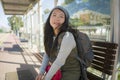 Outdoors portrait of young happy and beautiful Asian Korean tourist woman with backpack sitting cheerful on train station bench Royalty Free Stock Photo