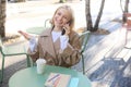 Outgoing young smiling woman, sitting in outdoor cafe, drinking coffee, answer phone call, talking on mobile, chatting