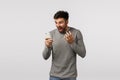 Outraged and furious pissed, tensed bearded man in grey sweater, holding smartphone, cursing, swearing at mobile display Royalty Free Stock Photo