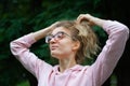 Outside portrait of blonde girl with hipster eyeglasses in pink clothes making ponytail outdoors in the urban park
