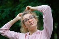 Outside portrait of blonde girl with hipster eyeglasses in pink clothes making ponytail outdoors in the urban park