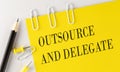 OUTSOURCE AND DELEGATE word on yellow paper with office tools on white background