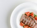 Oven-barbecued pork steak cut in slices, served in a white plate with fresh tomatoes and raw greenery.