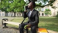 Overburdened African American young man in suit sitting on bench talking on phone and on tablet. Portrait of busy