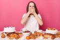 Overeating sad brown haired woman in white t shirt sitting at table isolated over pink background feeling nausea after eating