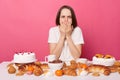 Overeating sick woman wearing white T-shirt sitting at festive table with various desserts, isolated over pink background,