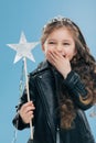 Overemotive positive small child covers mouth with one palm, wears fashionable black leather jacket and crown, holds magic wand in