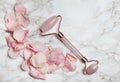 Overhead shot of a pink facial roller next to pink rose petals on a white marble surface