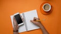 Overhead shot of stylish holding mobile phone and making note in notebook on orange background.