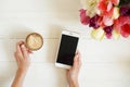 Overhead shot of woman hands holding cell phone gadget & coffee cup w cappuccino latte art on white wooden table background. Beaut Royalty Free Stock Photo