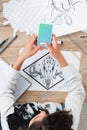 overhead view of female designer using smartphone with twitter application on screen at working table