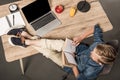 overhead view of schoolboy reading book and holding legs on table with laptop textbook lamp clock apple and glass of Royalty Free Stock Photo