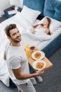 Overhead view of smiling man holding wooden tray with breakfast while woman sleeping Royalty Free Stock Photo