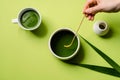 Overhead of a woman`s hand mixing with a matcha spoon in a bowl standing next to a cup