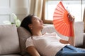 Overheated exhausted Asian woman waving paper fan, sitting on couch