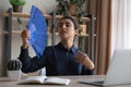 Overheated tired Indian businesswoman waving paper fan at workplace