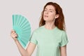 Overheated young red-haired woman using fan cooling herself.