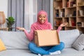 Overjoyed african american muslim lady opening cardboard box, receiving online shop order at home, sitting on sofa Royalty Free Stock Photo