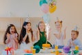Overjoyed children friends clap hands and looks at big sparkle on cake, stand near festive table with present, cups and holiday