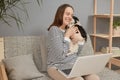 Overjoyed cute charming smiling Caucasian woman wearing striped shirt sitting on sofa with her puppy dog posing in front of laptop Royalty Free Stock Photo