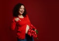 Happy ethnic pregnant woman with a bouquet of red tulips, pointing her index finger at her bare belly, on red background Royalty Free Stock Photo