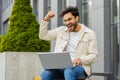 Happy Indian man working on laptop celebrate success win money get online good news outdoors street Royalty Free Stock Photo