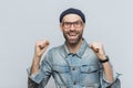 Overjoyed male rejoices his success, clenches fists, looks joyfully into camera, isolated over grey background. Bearded hipster g