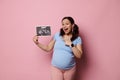 Overjoyed lovely pregnant woman points aside the ultrasound scan image of her future baby, isolated on pink background, Royalty Free Stock Photo