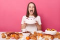 Overjoyed screaming brown haired woman in white t shirt sitting at table showing tasty cake breaking diet or having cheat meal