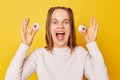 Overjoyed screaming young girl with ponytails in jumper showing sugary delicious marshmallow posing isolated over yellow Royalty Free Stock Photo