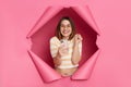 Overjoyed smiling happy positive brunette woman wearing striped t shirt posing in torn pink paper wall using smart phone and