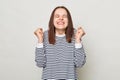 Overjoyed smiling woman with brown hair wearing striped shirt standing isolated over gray background keeps eyes closed clenched