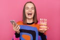 Overjoyed woman holding delicious appetizing doughnuts with burning candles wearing casual jumper using cell phone, standing