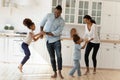 Happy biracial parents dance with little children in kitchen