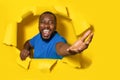 Overjoyed young black man stretching open palm through hole in torn yellow paper background, taking or giving something Royalty Free Stock Photo