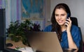 Overjoyed young woman in denim shirt working home with a laptop, talking via phone and looks to the laptop. World map in Royalty Free Stock Photo