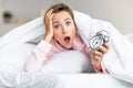 Overleeping. Portrait of shocked woman holding clock in bed