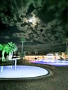 Pools of water illuminated with blue light surrounded by palm trees against a dark sky with a moon and beautiful clouds Royalty Free Stock Photo