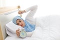 Oversleeping asian muslim woman wearing white sleepwear lying on bed, missing ring of alarm clock waking up.