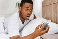 Overslept African Man Holding Alarm-Clock Waking Up In Bed Indoor