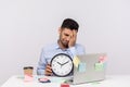 Overtime work. Unhappy sorrowful man employee sitting in office workplace, holding clock and keeping facepalm Royalty Free Stock Photo