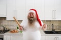 An overweigh man in Santa Claus hat with tattooed arms wearing a white T-Shirt, holding a wooden rolling pin on one hand and