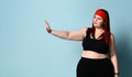 Overweight ginger girl in red headband, black top, leggings, sneakers. Showing stop sign by her palm, posing on blue background