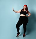 Overweight ginger girl in red headband, black top, leggings, sneakers. Showing stop sign by her palm, posing on blue background