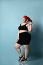 Overweight ginger lady in red headband, black top, shorts, sneakers. Smiling, touching her ponytail, posing on blue background