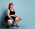 Overweight ginger model in red headband, black top, shorts, sneakers. Squatting posing sideways against blue studio background